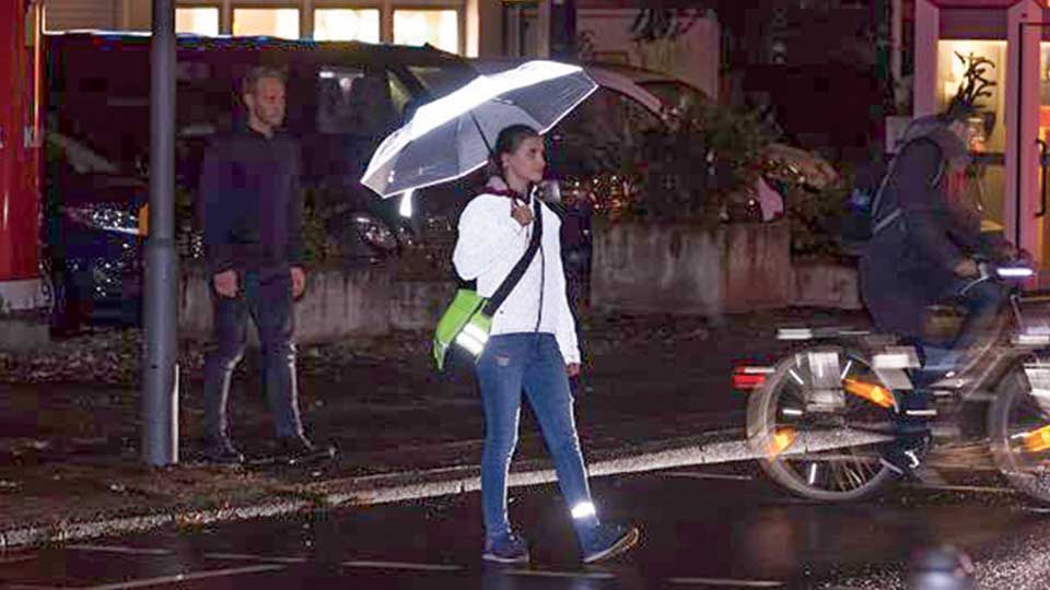 Straßenszene in der Dunkelheit. Eine Frau mit stark reflektierender Jacke, Schirm und Hosenband überquert angestrahlt und gut sichtbar eine Straße.