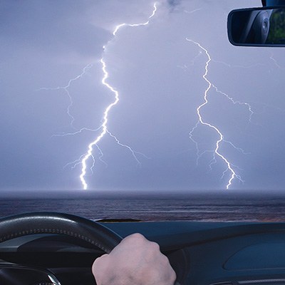 Blick aus einem Auto über das Lenkrad, das eine Hand hält, auf einen mit dunklen Wolken gefüllten Himmel, durch den gerade ein Blitz fährt.