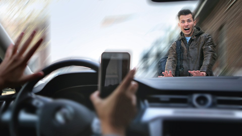 Blick aus einem Auto durch die Windschutzscheibe. Die Fahrerin hält ein Handy in der Hand und achtet nicht auf den Verkehr. Vor dem Auto steht ein erschrockener Passant und stützt die Hände auf der Motorhaube ab.