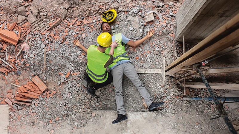 Ein junger Mann in Arbeitsbekleidung mit gelber Warnweste liegt offenbar abgestürzt bewusstlos auf dem steinigen Boden einer Baustelle. Ein Kollege kniet neben ihm und massiert sein Herz.