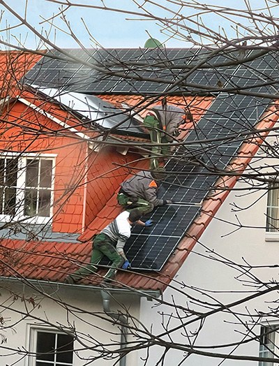 Blick auf ein Dach, auf dem drei Arbeiter an Photovoltaik-Elementen arbeiten. Es gibt keinerlei Absicherung gegen Absturz – eine extrem gefährliche Situation.