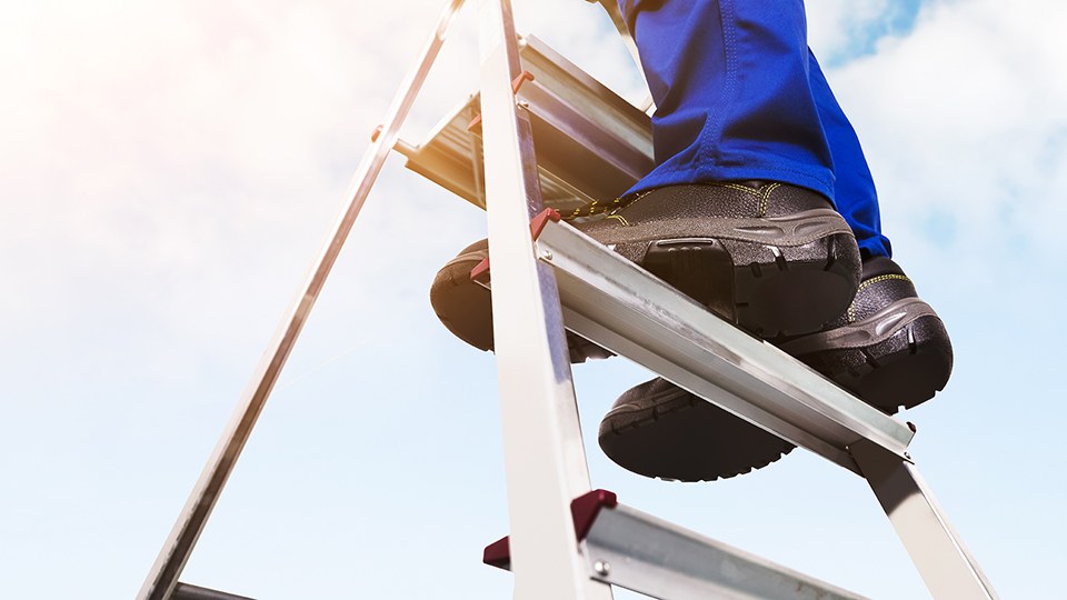 Blick von unten auf die Beine eines Handwerkers in blauer Arbeitshose und Sicherheitsschuhen. Er steht auf der vorletzten Trittfläche einer Klappleiter.