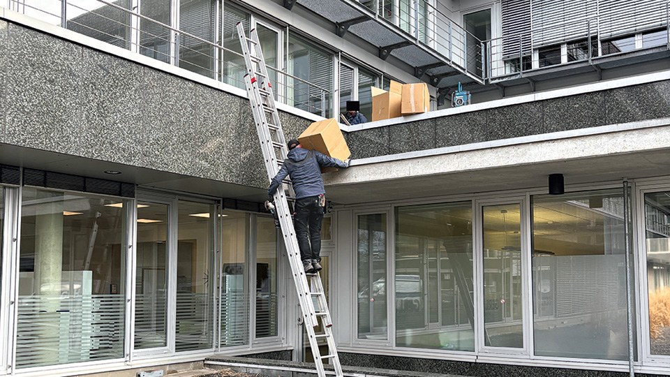 Ein Arbeiter klettert eine Anlegeleiter hoch. Dabei transportiert er ein großes Paket, das er kaum mit einem Arm halten kann, zum nächsten Gebäudestockwerk.