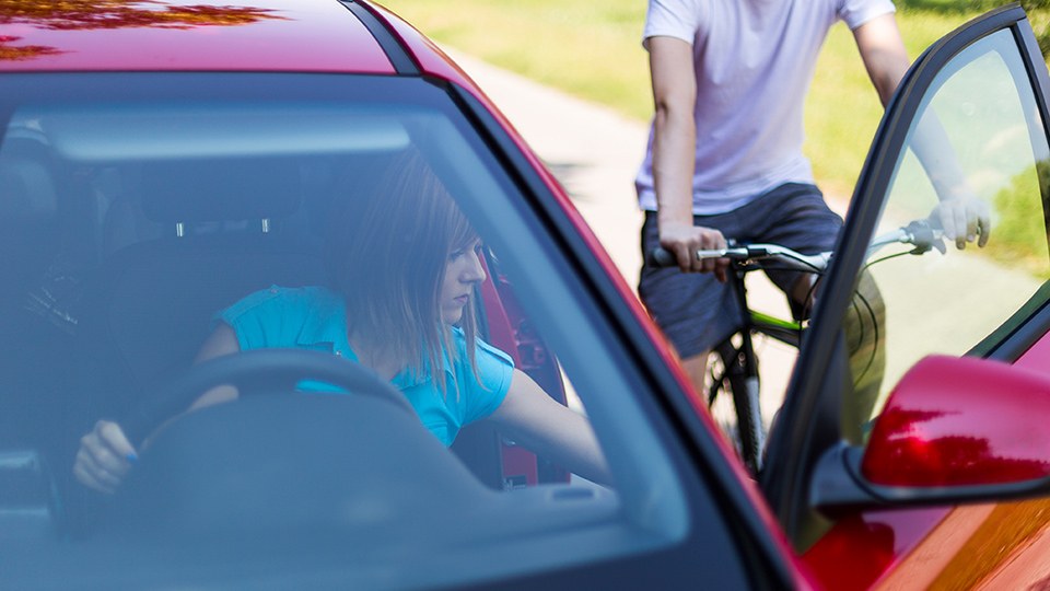Eine Autofahrerin öffnet ihre Fahrertür, von hinten nähert sich ein Fahrradfahrer, es wird zur Kollision kommen.