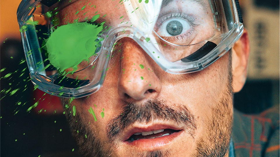 Mann mit Arbeitsschutzbrille, der die Augen aufreißt, als ihm eine grüne Flüssigkeit auf die Brille spritzt.