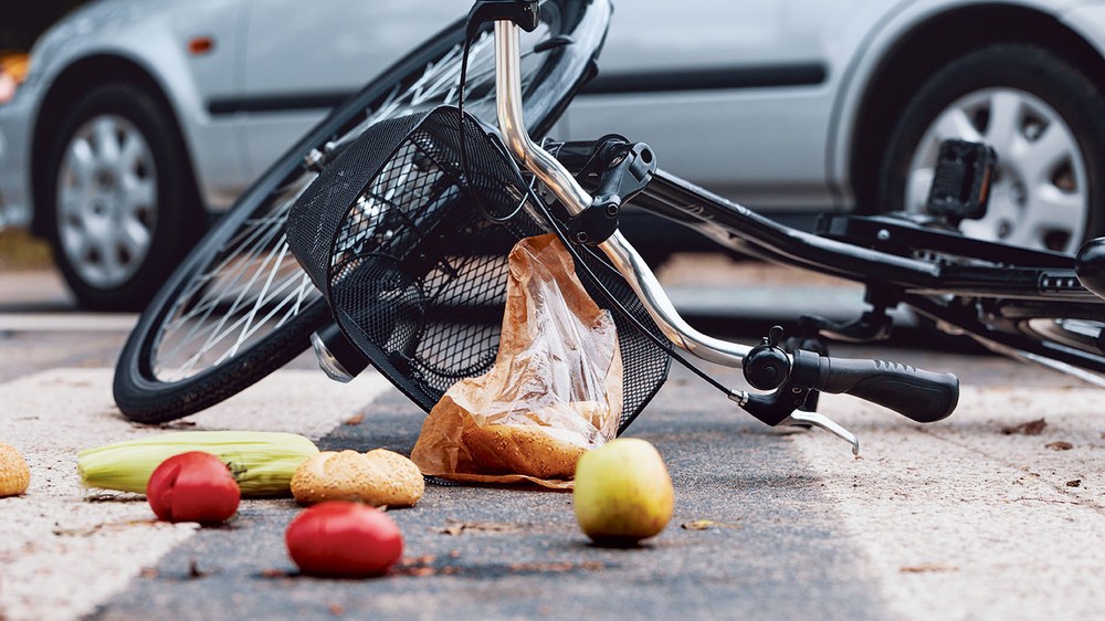 Ein Fahrrad, Obst und andere Einkäufe liegen nach einem Unfall auf der Straße, im Hintergrund ein silberner Pkw