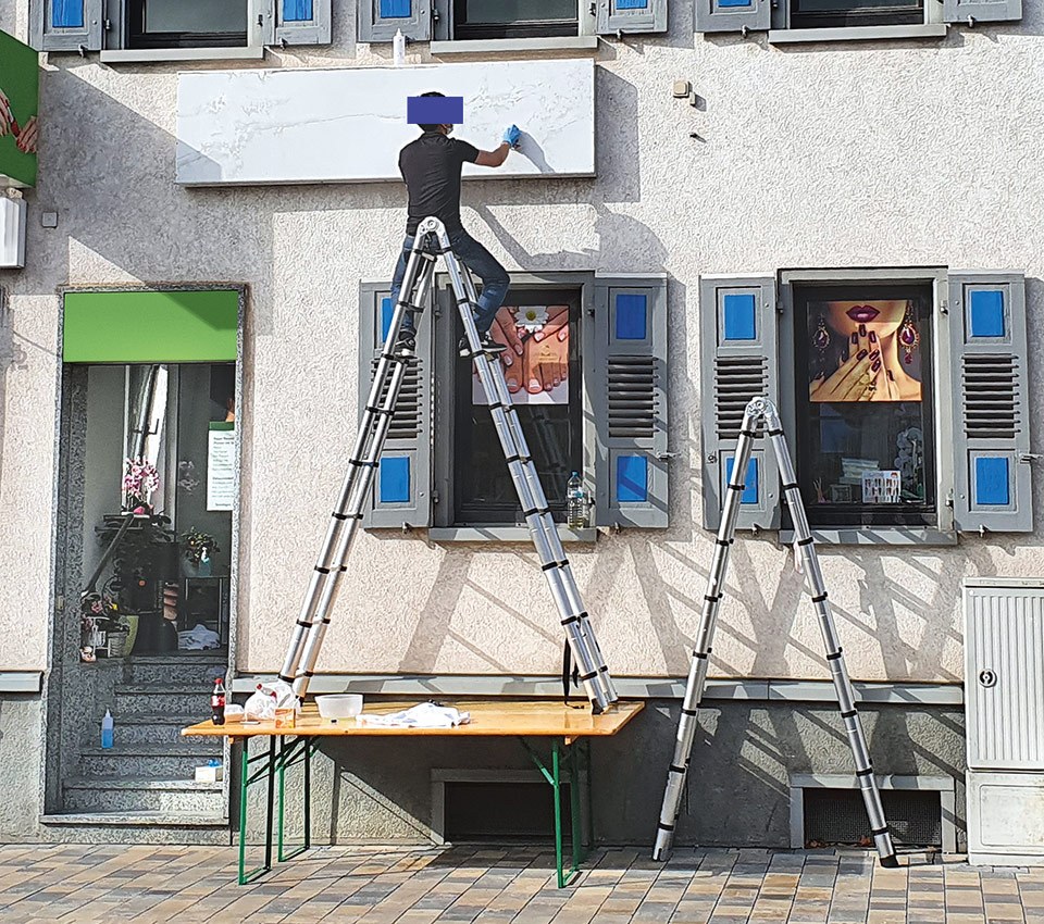 Ein Arbeiter bei Arbeiten an der Außenfassade eines mehrstöckigen Gebäudes. Er steht ungesichert auf einer Bockleiter, die wiederum auf einem Klapptisch steht.