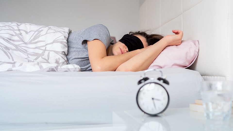 Schlafende Frau im Bett; der Raum ist hell, sie trägt eine schwarze Schlafmaske. Im Vordergrund ein Wecker, der 15:30 Uhr zeigt.