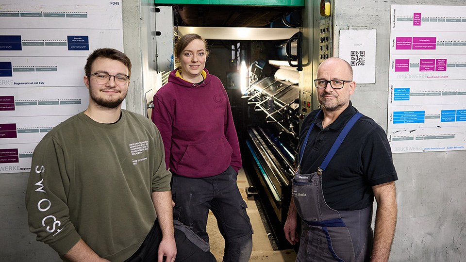 Altersgemischte Teams bei Mohn Media Niko Brinkmann, Tamara Wetzel und Markus Severins am Zugang zum Inneren einer riesigen Druckmaschine; sie tragen Arbeitshosen, Severins einen Overall, und blicken in die Kamera. Links und rechts von ihnen an der Wand Pläne zur Belegung der Druckmaschine.