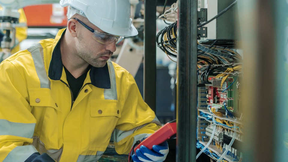Elektriker mit Helm, Schutzbrille, Handschuhen und Sicherheitsjacke prüft etwas an einer Schaltanlage