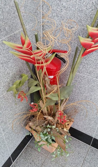 Ein roter Feuerlöscher, der in einer Ecke versteckt ist. Davor eine Vase mit Blumendeko, die den Feuerlöscher verdeckt.